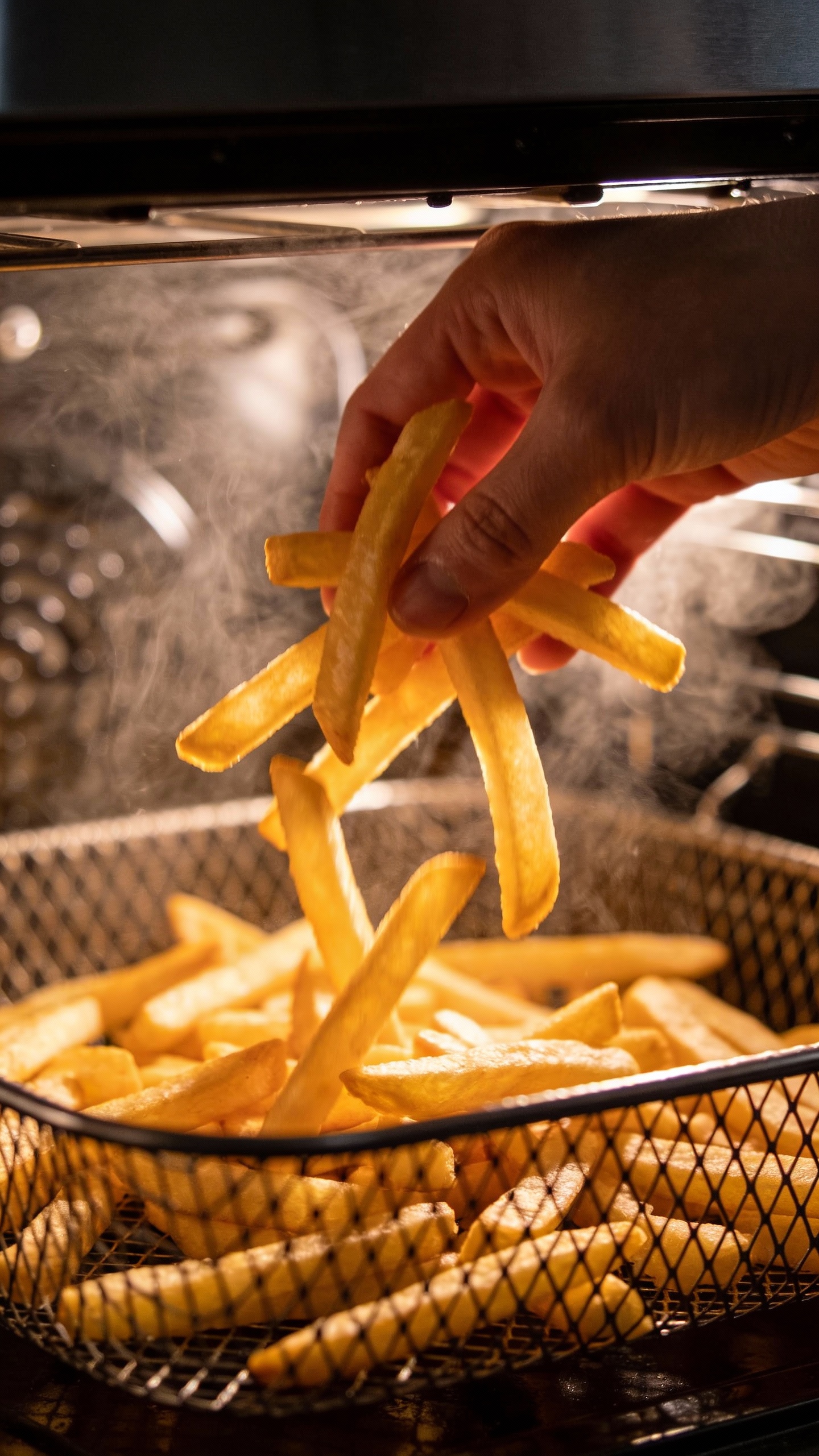 hand tossing fries mid-cook in air fryer basket, steam